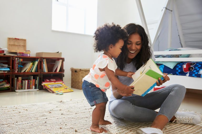 Woman sitting cross legged on the floor, smiling and showing a book to the toddler standing next to her