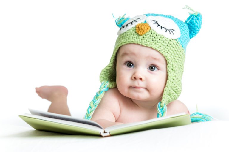 baby in a crocheted hat lying on its stomach in front of an open book