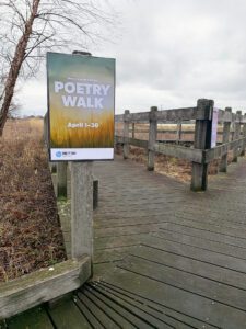 A photo of the boardwalk outside the library, with a sign that says "Poetry Walk, April 1-30"