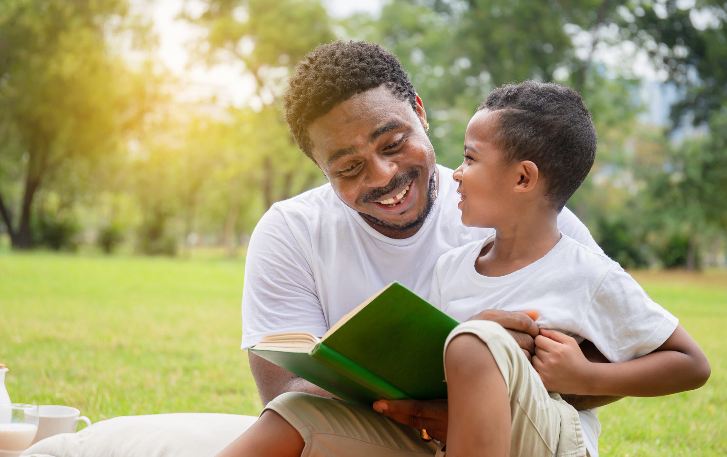 Cheerful AfricanAmerican boy and dad reading in the park
