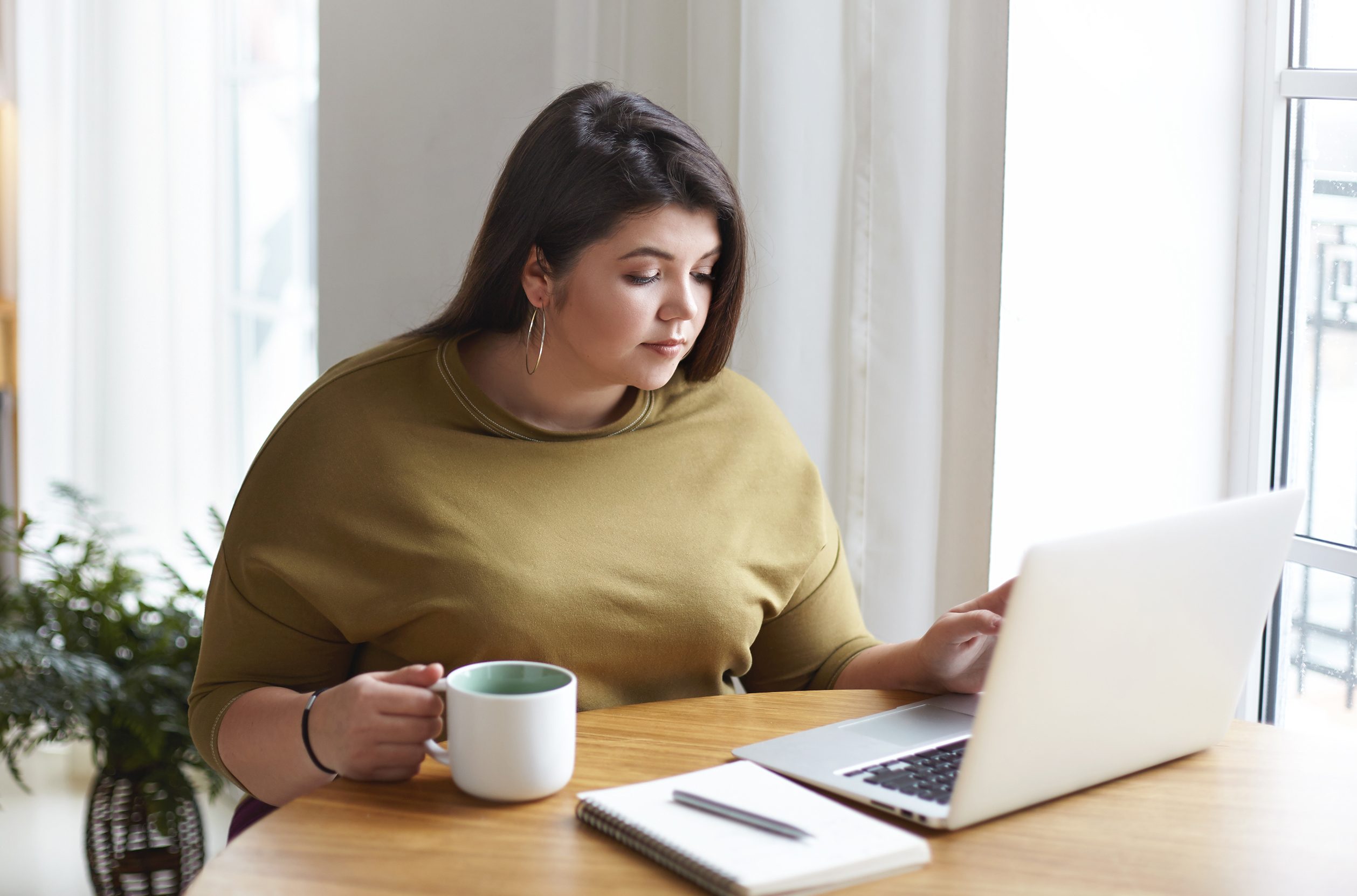 Relaxed woman sitting at table with coffee mug and laptop, reading the computer screen