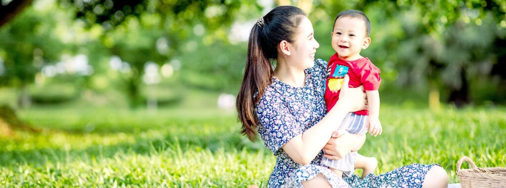 Smiling woman holding a happy baby while seated outside on the grass.