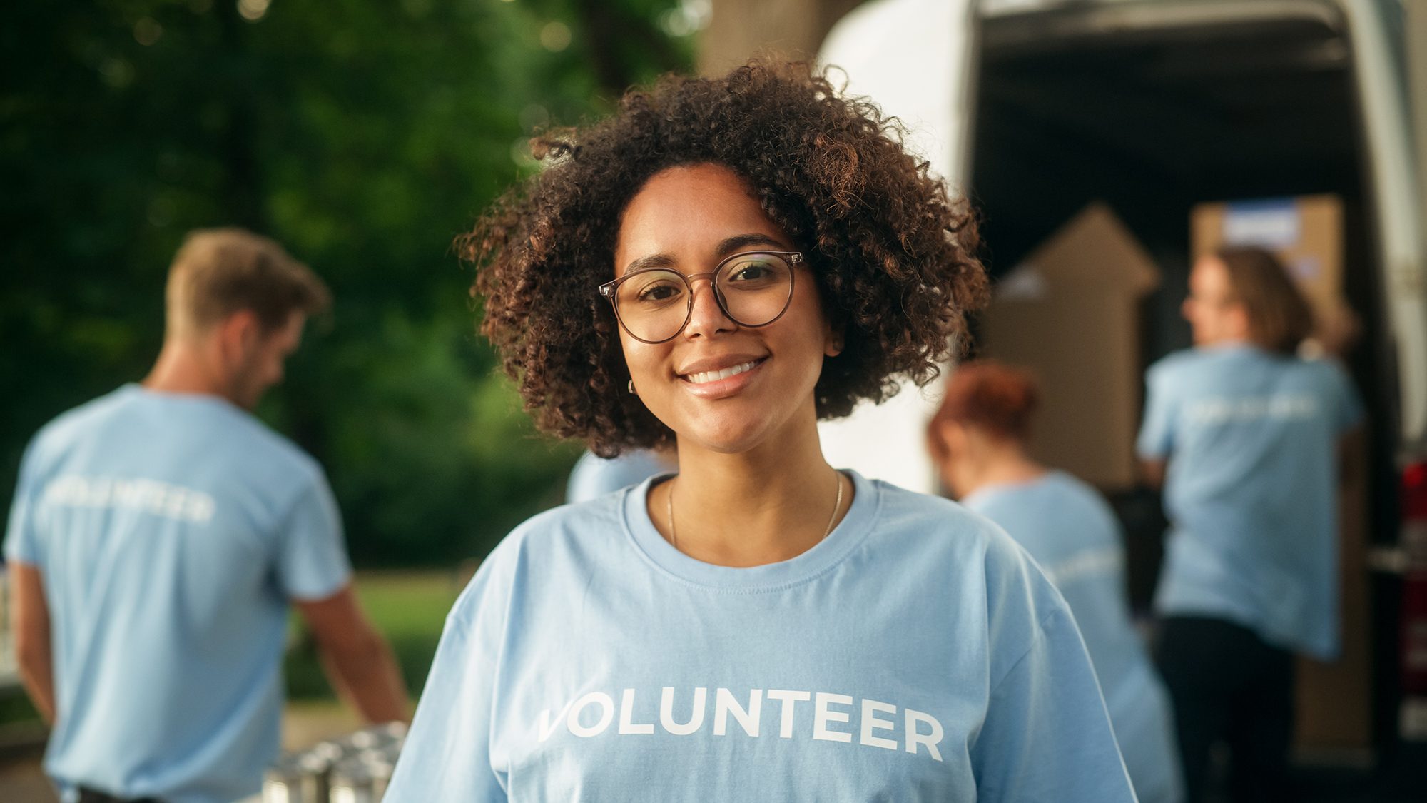 teenager smiling and wearing volunteer t-shirt while other teens are volunteering behind her.