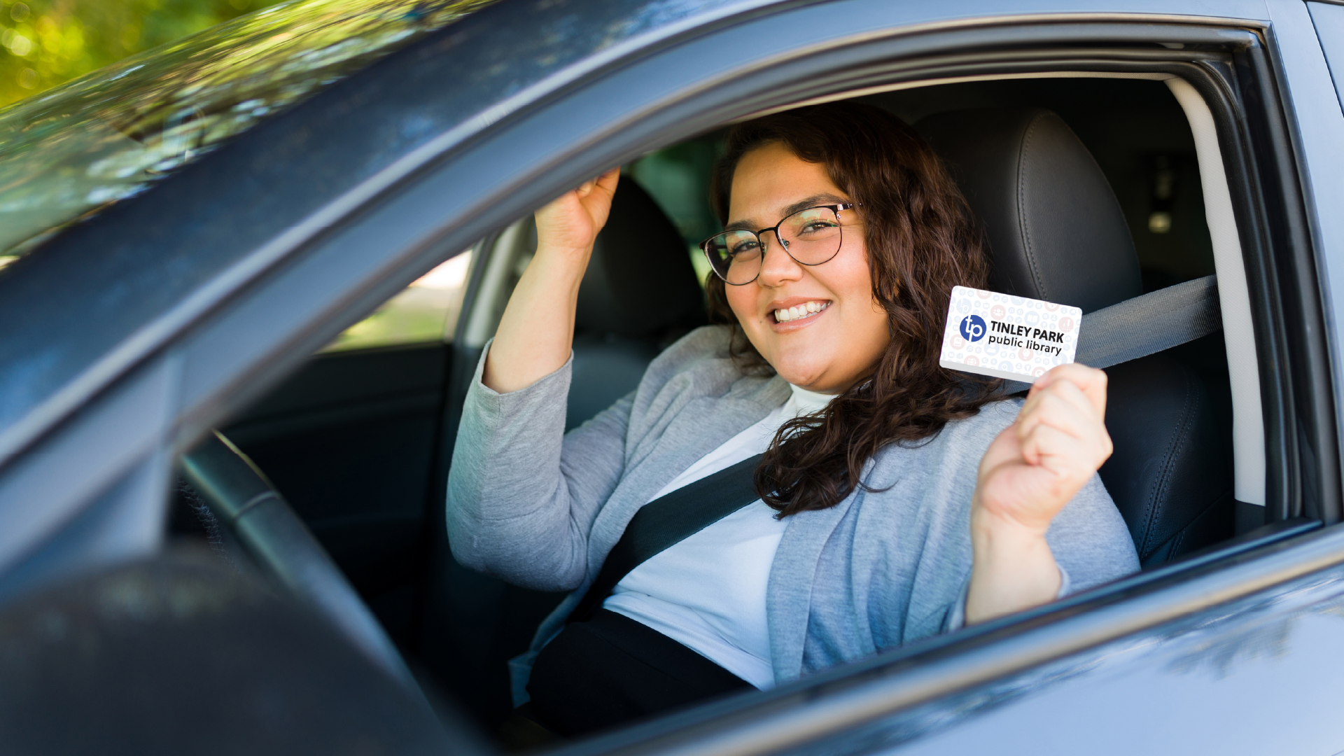 Woman in driver's seat of car with window rolled down, smiling and holding a library card