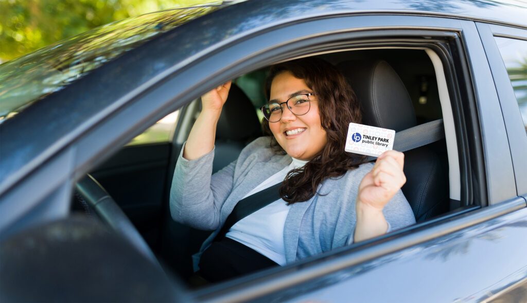 Woman in driver's seat of car with window rolled down, smiling and holding a library card