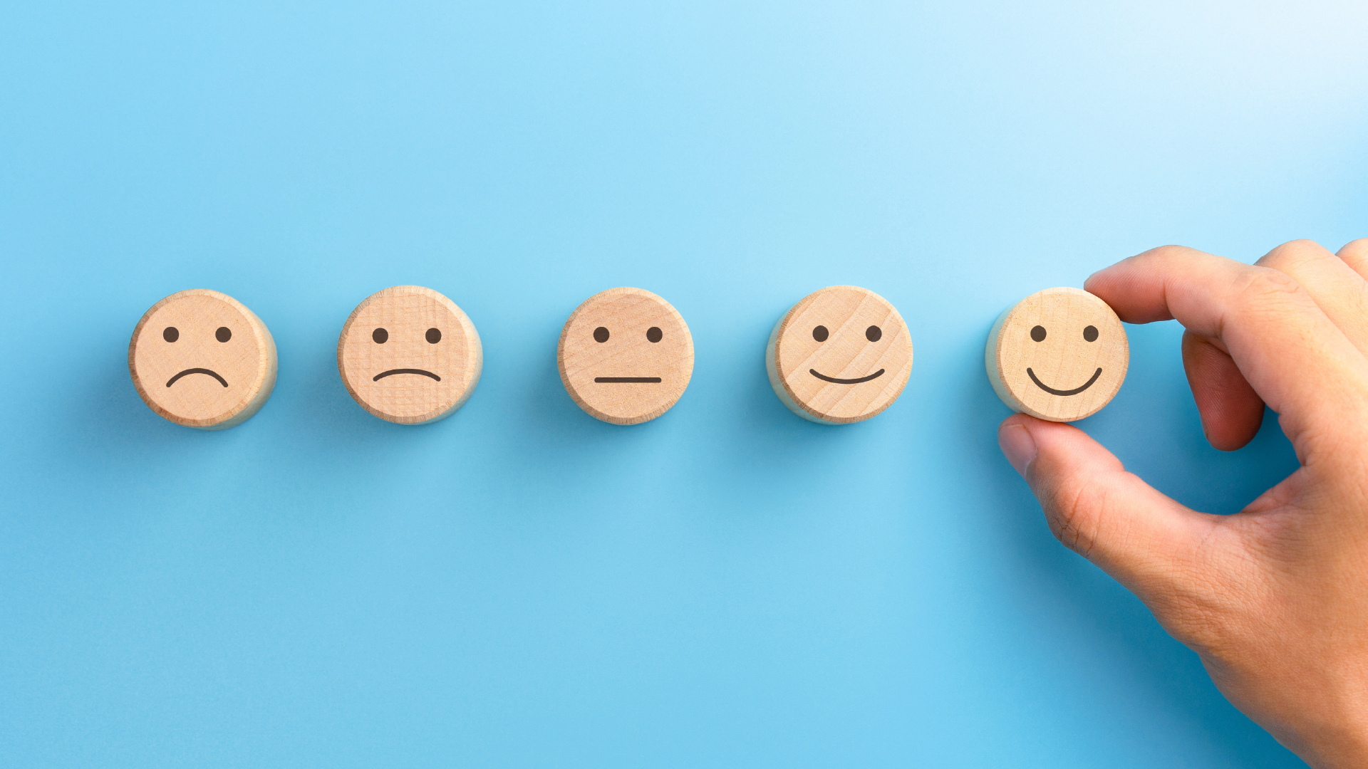 A row of five wood-carved smiley faces, arranged from sad to happy. A hand is holding the most happy smiley face.